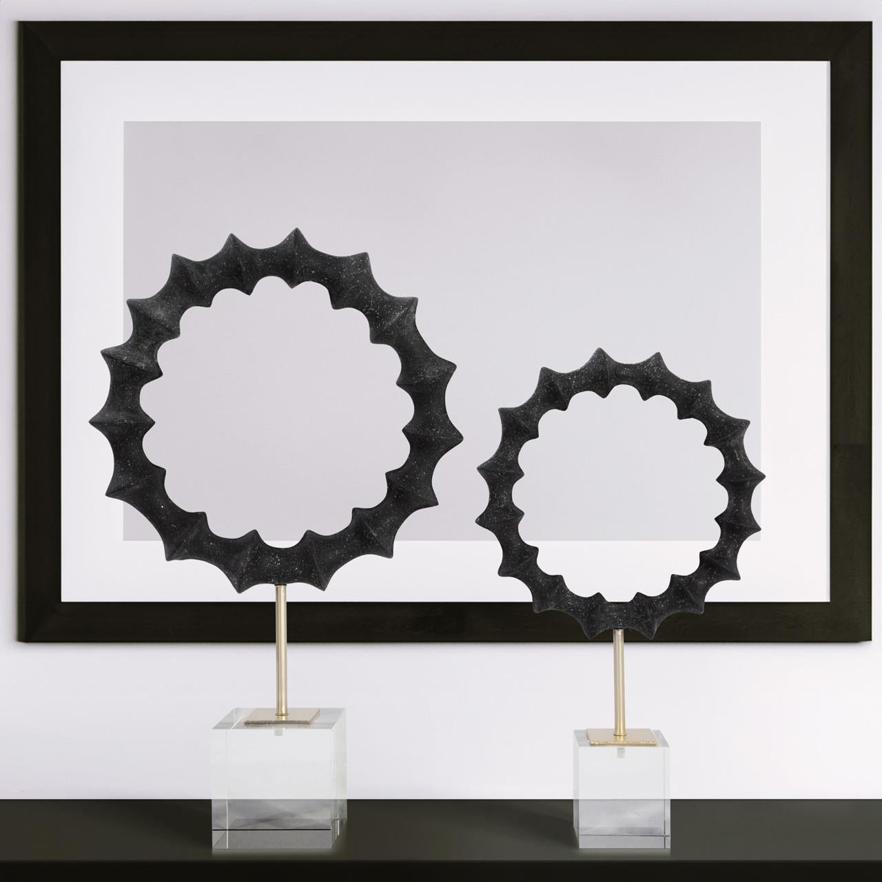 Two abstract sculptures on stands with a black and white framed print background.