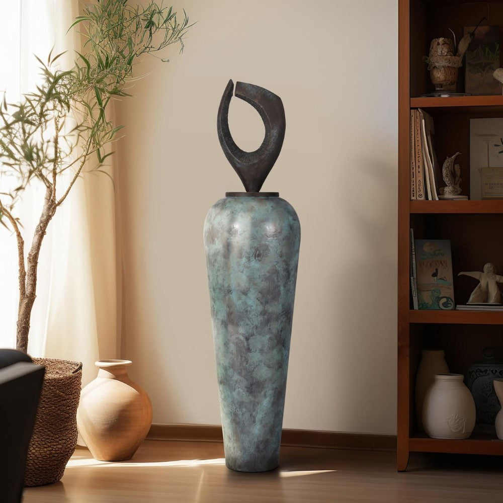 Tall vase with abstract top, next to an olive branch and shelf in a sunlit room.