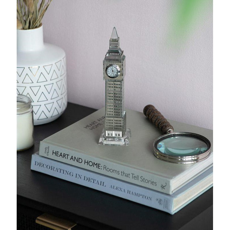 Mini clock tower on books with vase and magnifying glass.