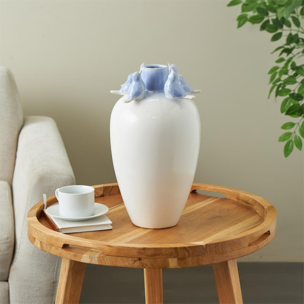 White vase with blue birds on table, beside a sofa and cup.