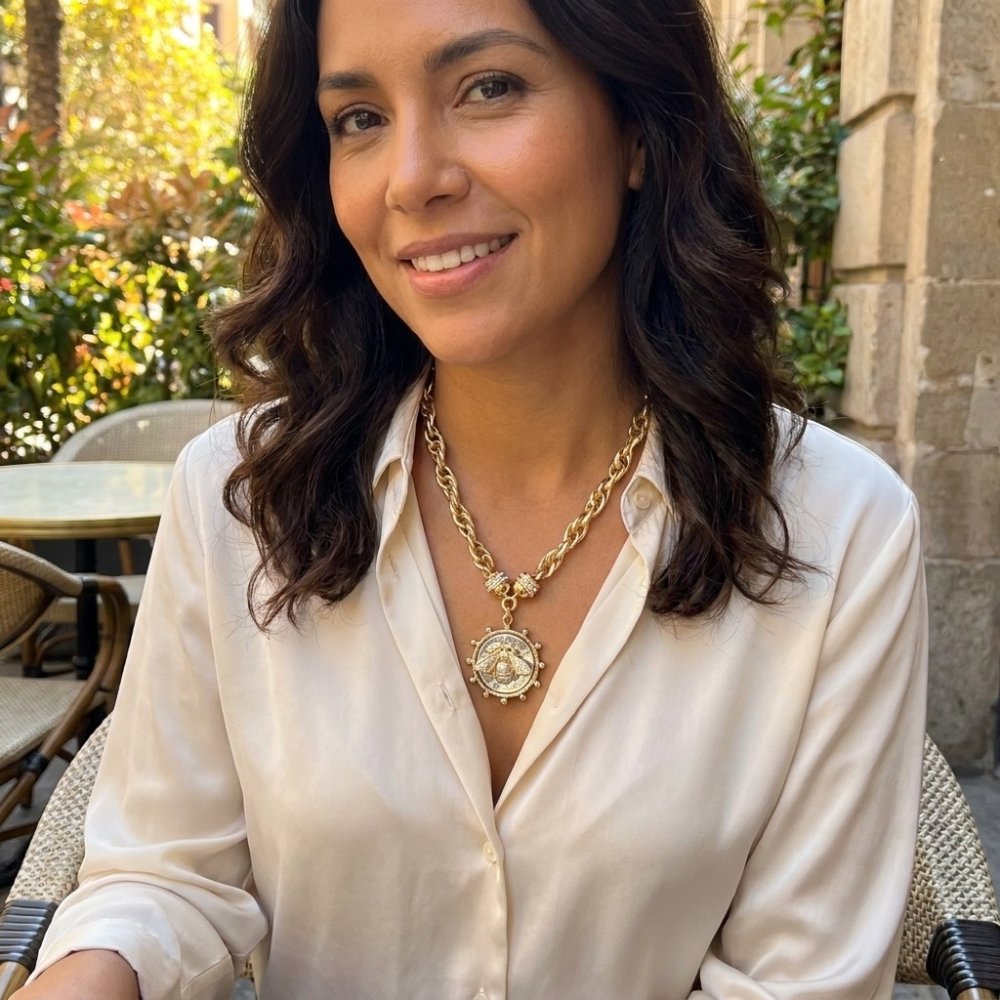 A woman smiles outdoors in a white blouse and a gold pendant necklace.