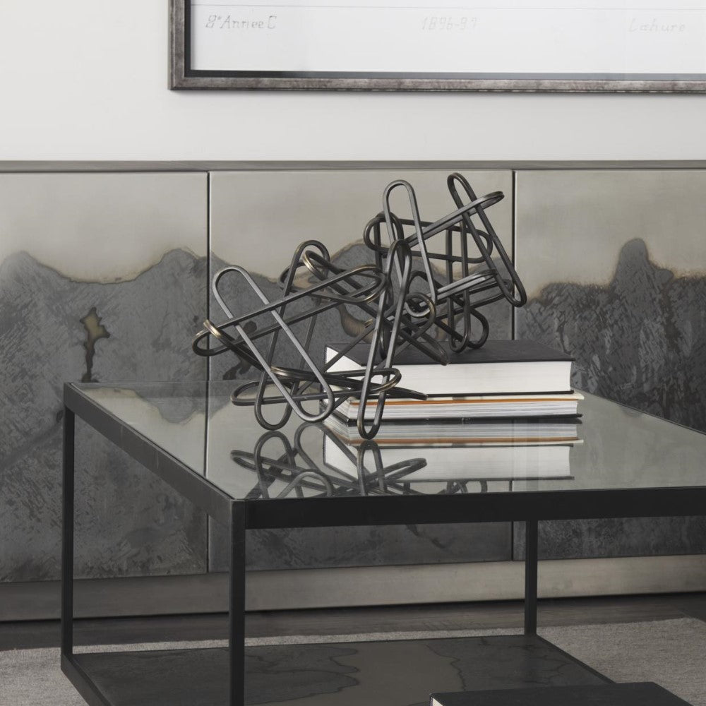 Modern metal sculpture on a glass table with books and abstract art in the background.