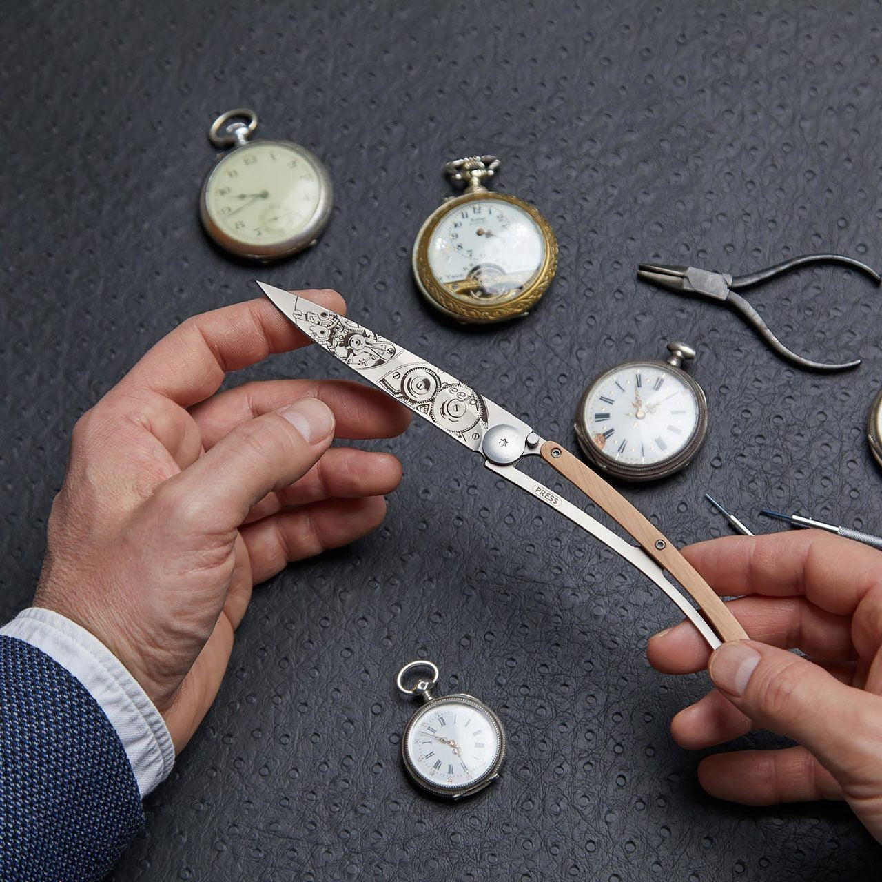 Hands holding ornate knife with pocket watches on textured surface.
