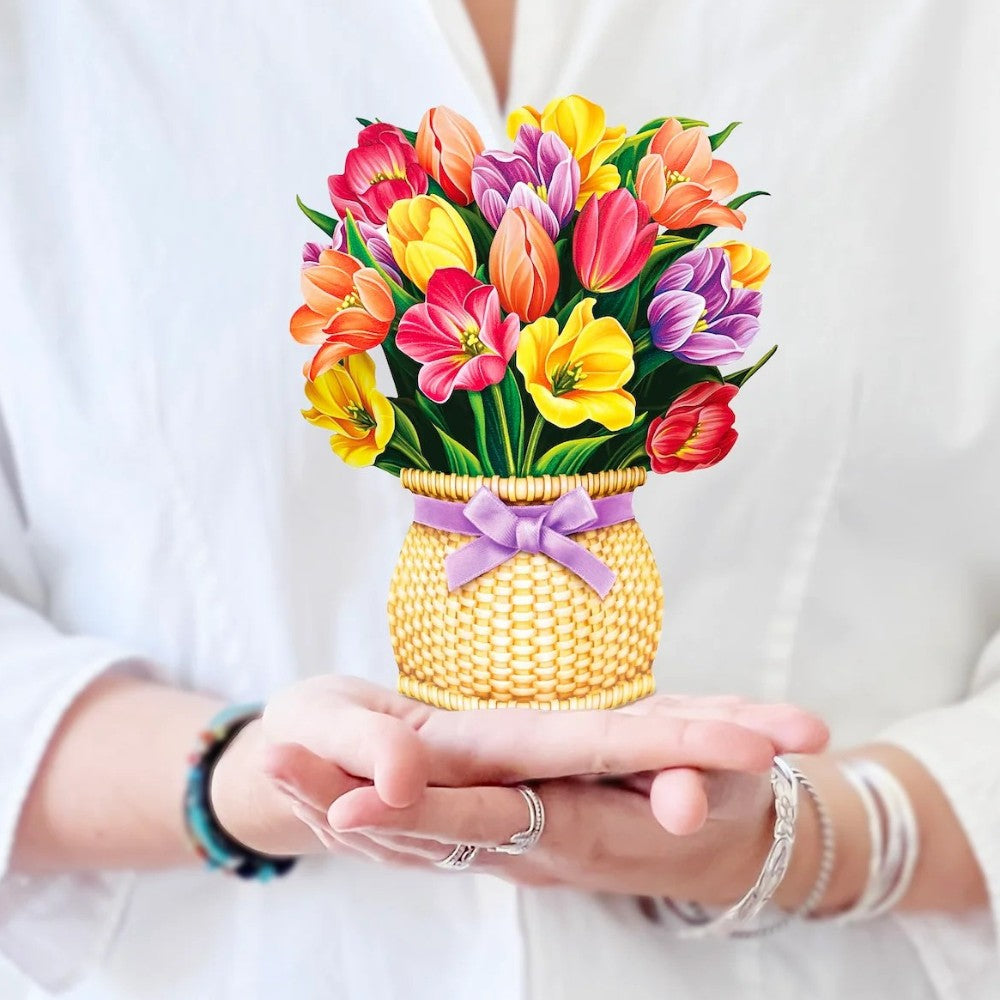 Person holding a basket of colorful flowers adorned with a purple bow.