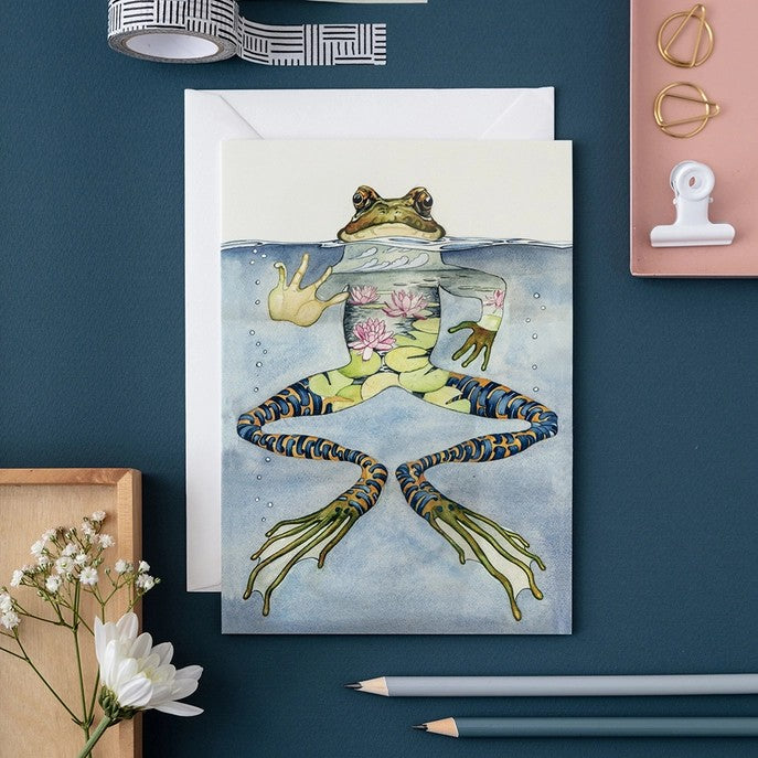 Artistic frog card on a desk with flowers and stationery.