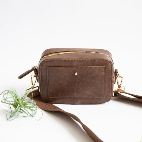 Brown leather crossbody bag with gold zippers and strap, next to a small plant.