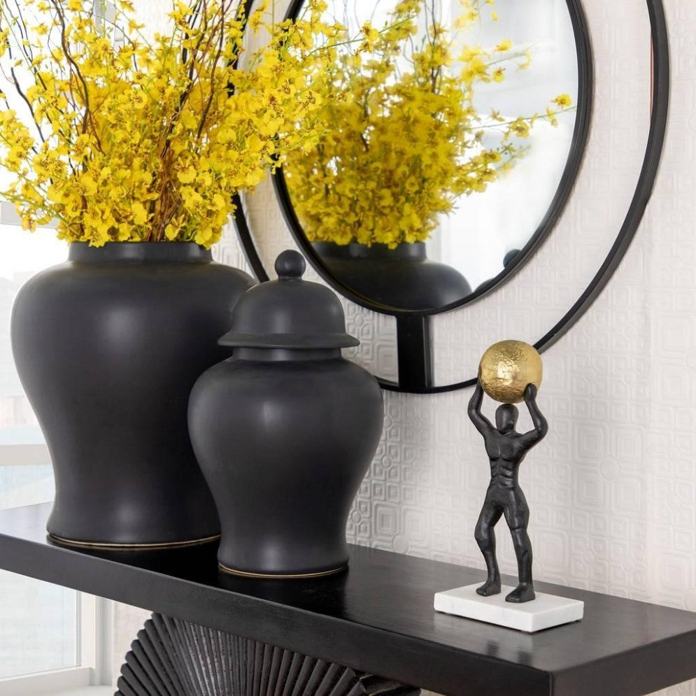 Black vases, yellow flowers, statue, and round mirror on a dark shelf.