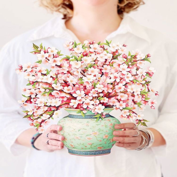 Person holding a vase filled with pink and white cherry blossoms.