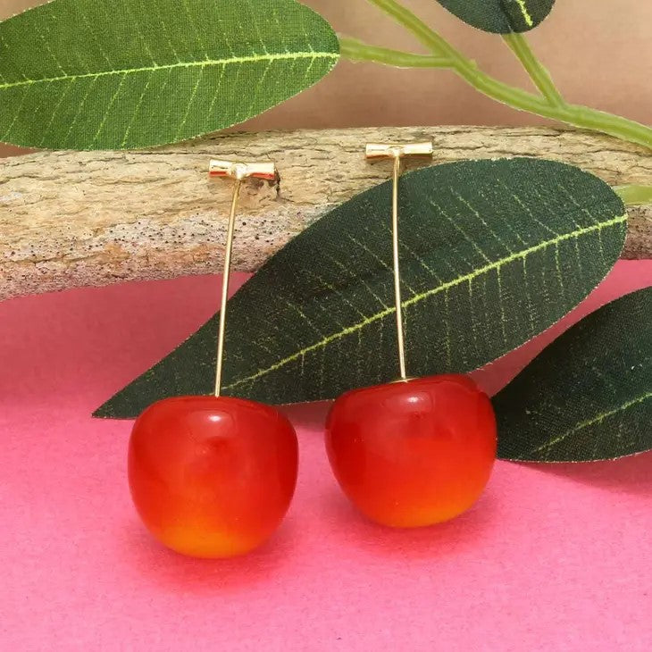 Red cherry earrings with green leaves on pink background.