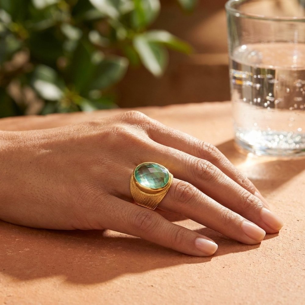 A hand wearing a gold ring with a large green gemstone rests on a sunlit table.