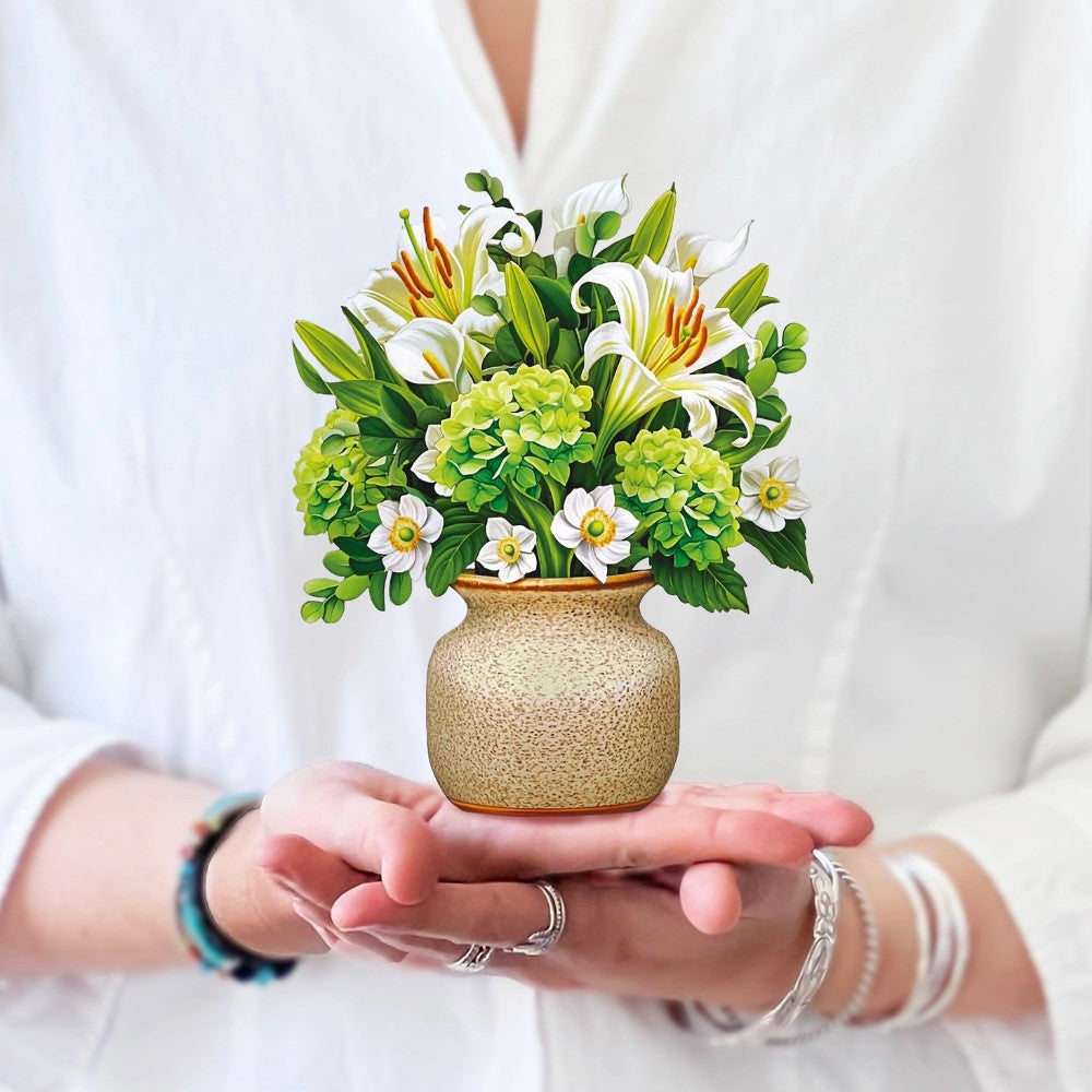 Hands holding a tiny flower pot with white lilies and green blooms.