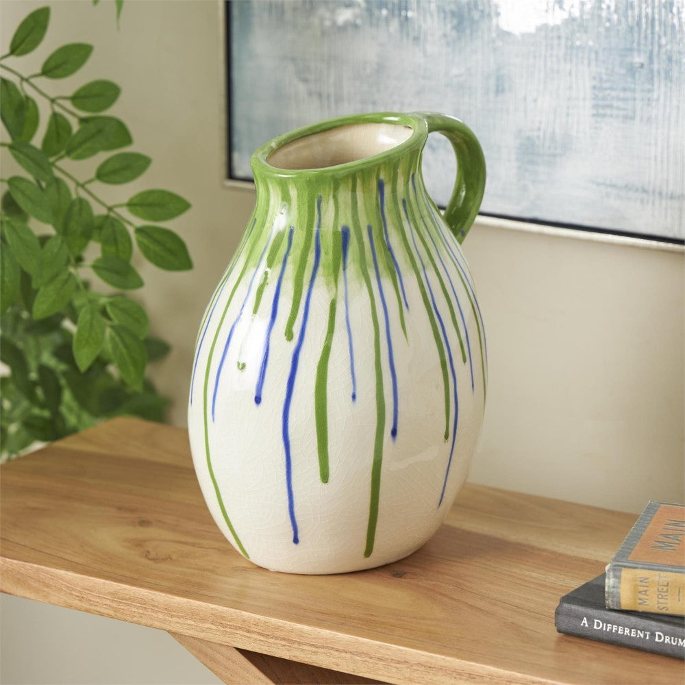 Ceramic jug with green and blue drips on a wooden table next to books and a plant.