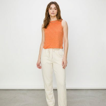 Woman in orange top and cream pants against a white background.
