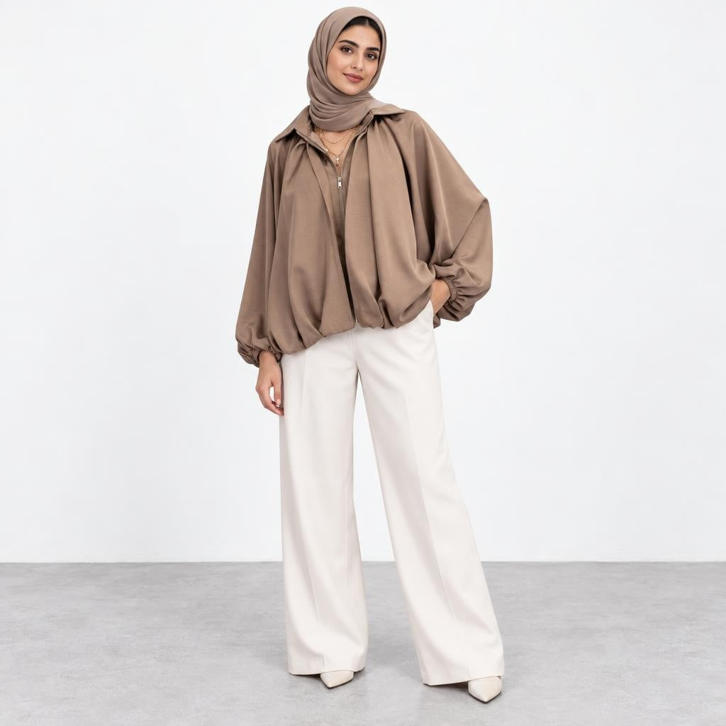A woman in a taupe hijab and jacket poses in wide-leg white trousers.