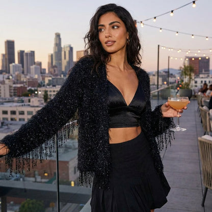 Woman in black outfit holds cocktail on city rooftop at sunset.