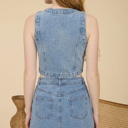 Person in a denim vest and skirt stands against a neutral background.
