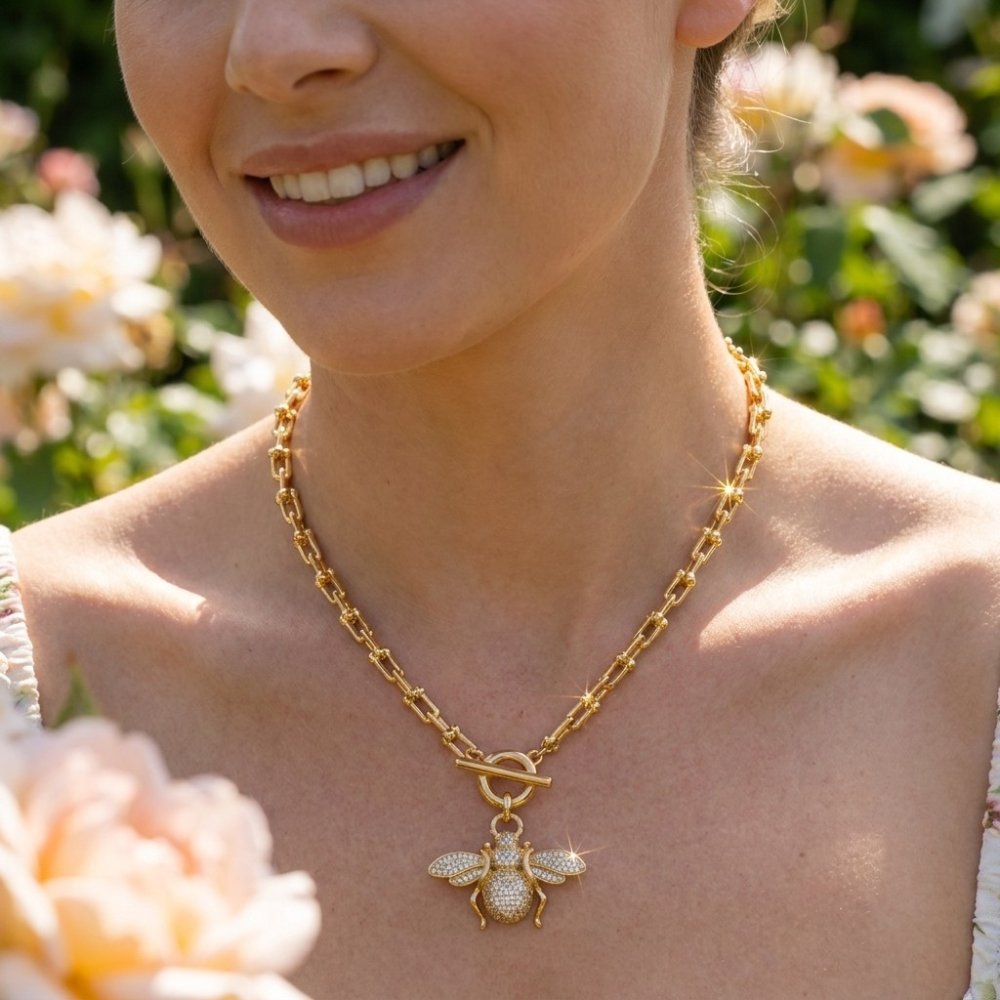 A smiling woman wears a gold bee pendant necklace among blooming roses.