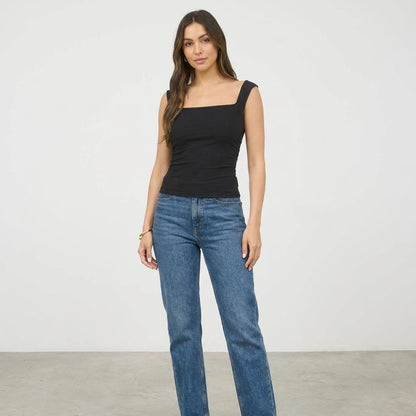 Woman in a black top and jeans stands against a plain background.