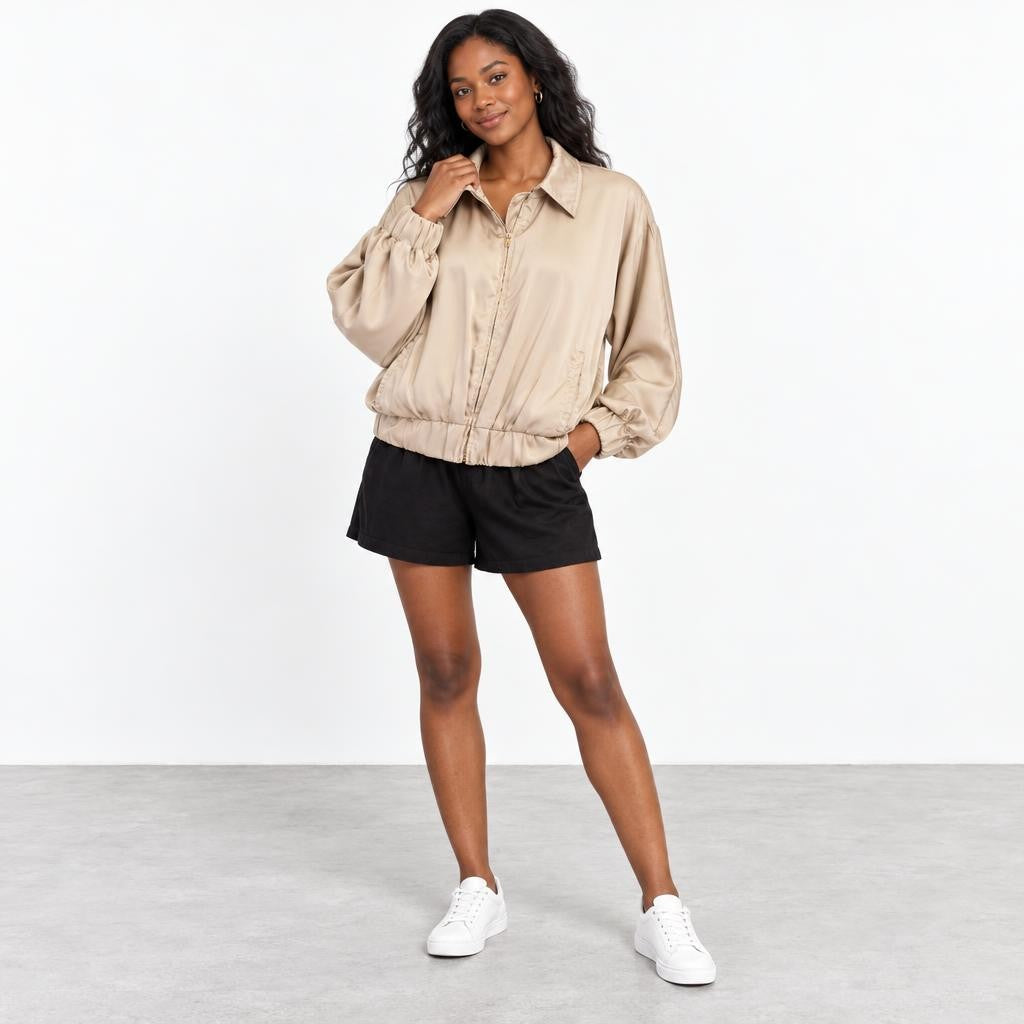 A woman models a beige jacket, black shorts, and white sneakers against a simple studio backdrop.
