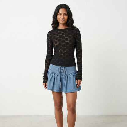 Woman in a black lace top and denim skirt stands against a light background.