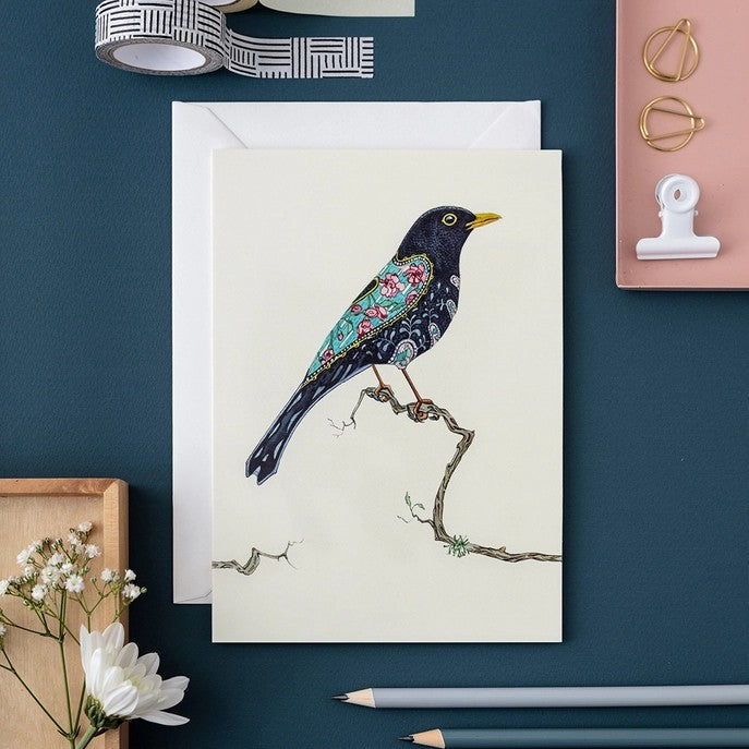 Decorative bird card on dark desk with stationery and flowers.