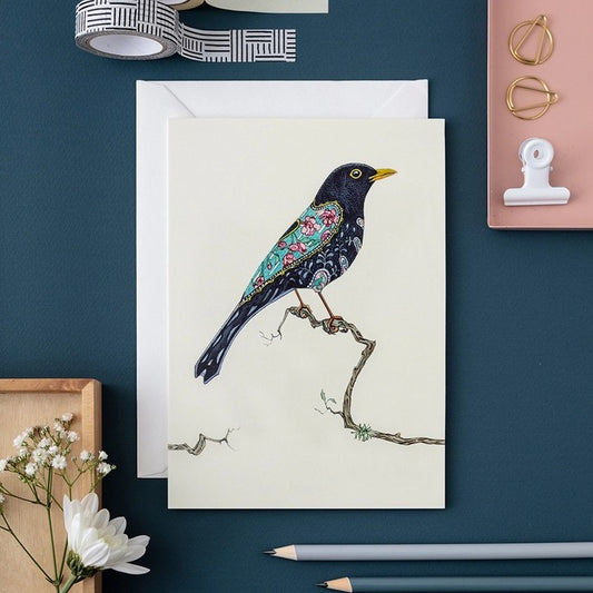 Decorative bird card on dark desk with stationery and flowers.