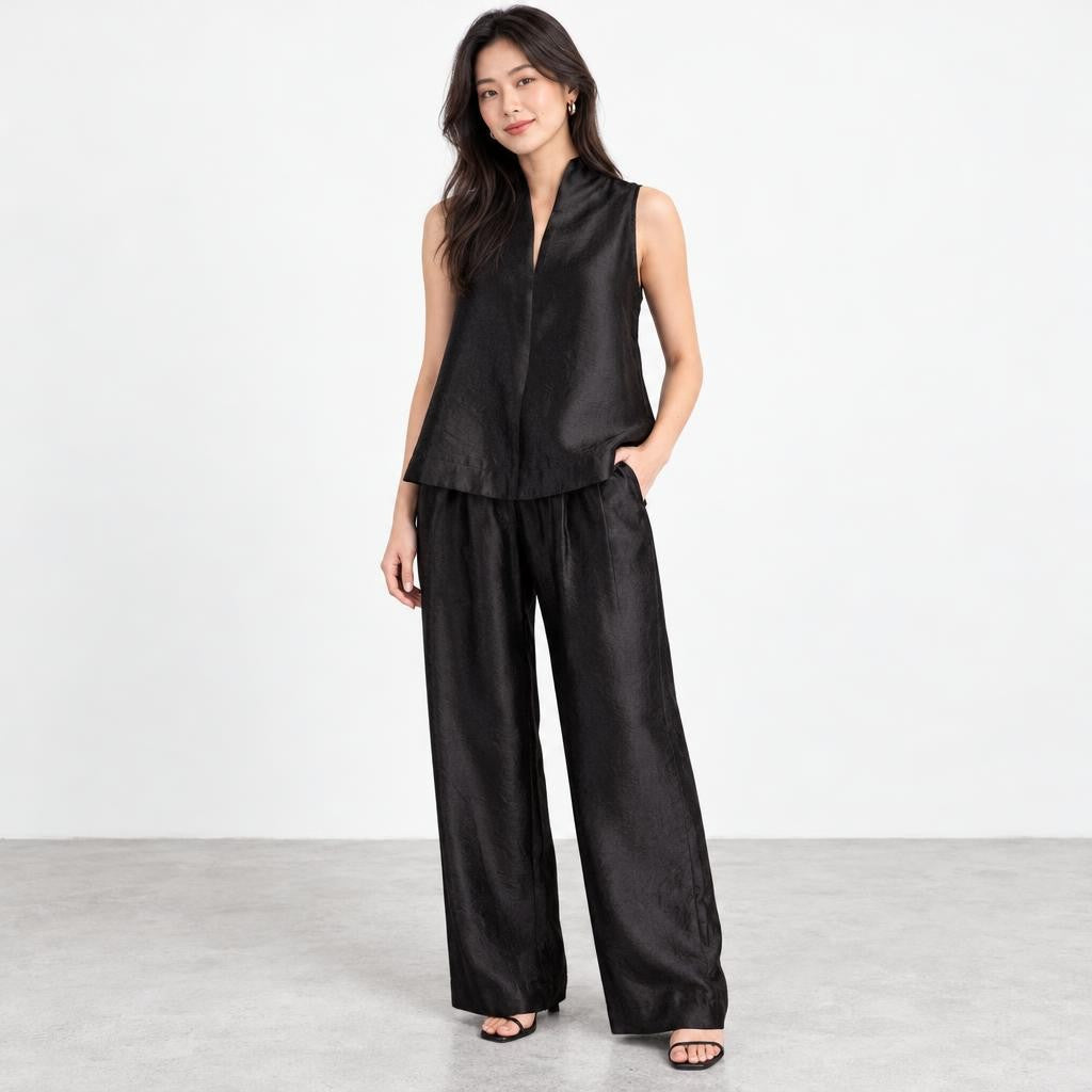 A woman models a sleek black sleeveless top and wide-leg trousers against a minimalist backdrop.