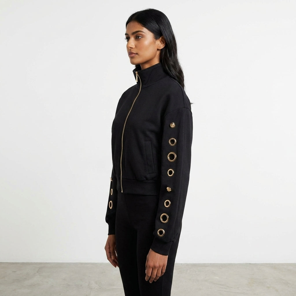 Woman in black jacket with gold accents stands in profile against plain background.