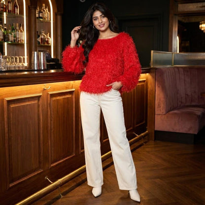 A woman in a bright red sweater and white pants poses beside a wooden bar.