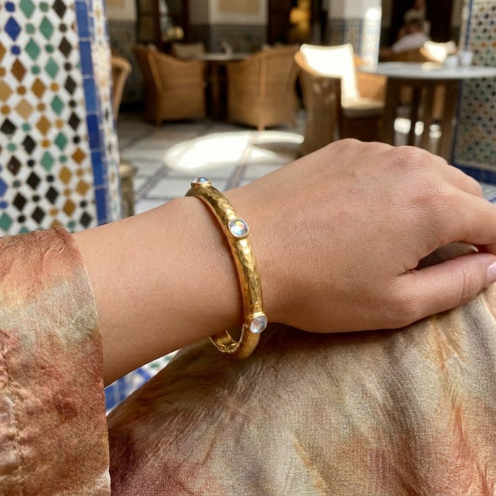 A hand wearing a gold bracelet with iridescent stones rests in a sunlit café.
