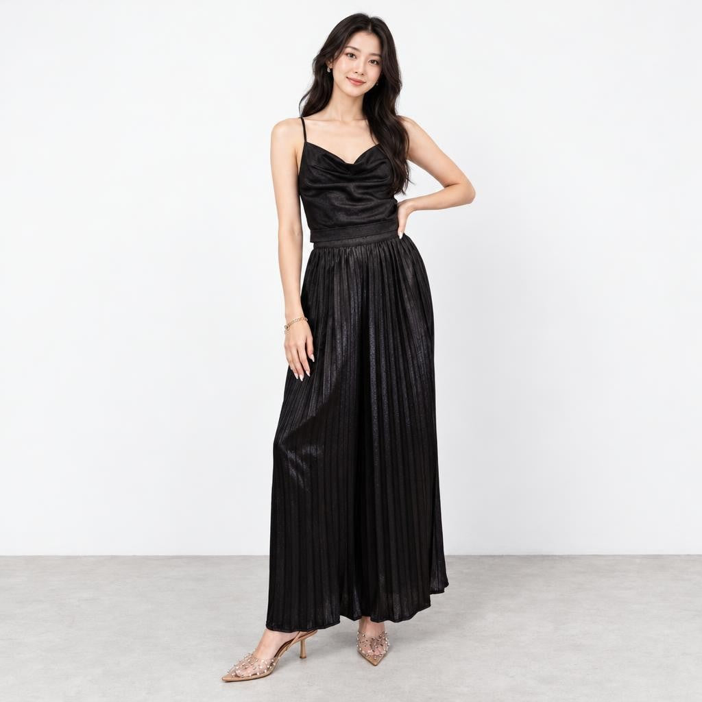 A woman poses in a black pleated maxi dress and embellished heels against a plain studio backdrop.