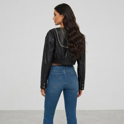 A woman models a cropped black leather jacket with gold detailing and blue jeans.