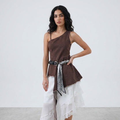 Woman in asymmetrical brown top, lace skirt, and belt stands against a plain backdrop.