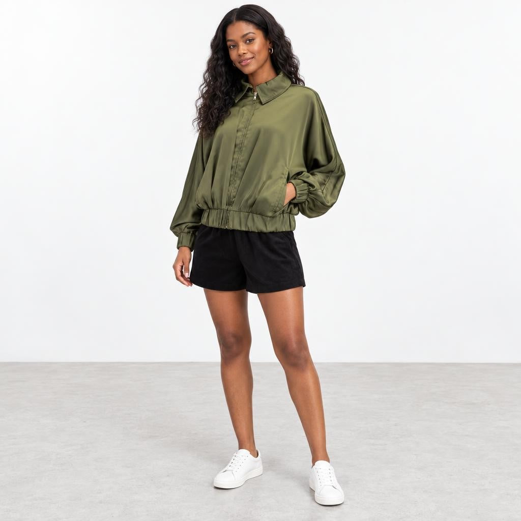 A woman models an olive-green satin jacket, black shorts, and white sneakers in a studio.