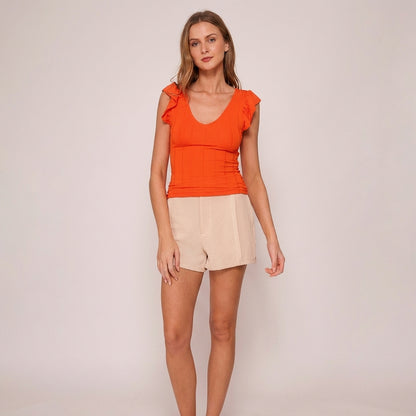 Woman in orange top and beige shorts against a plain background.