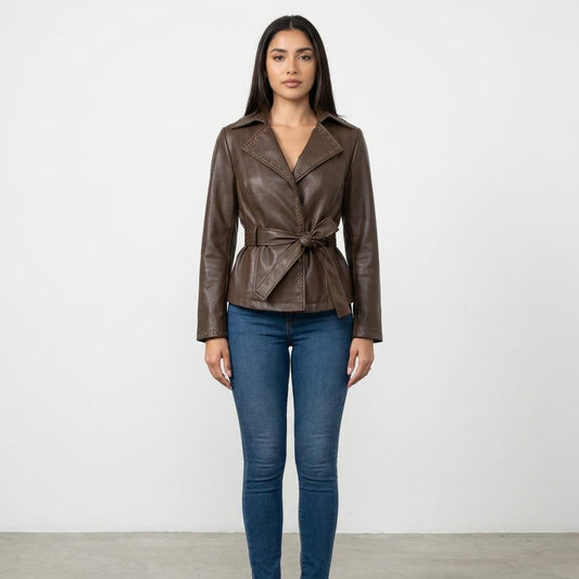 A woman models a belted brown leather jacket with blue jeans against a plain backdrop.