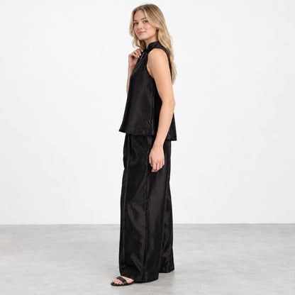 A woman models a black sleeveless top and wide-leg trousers against a plain studio backdrop.