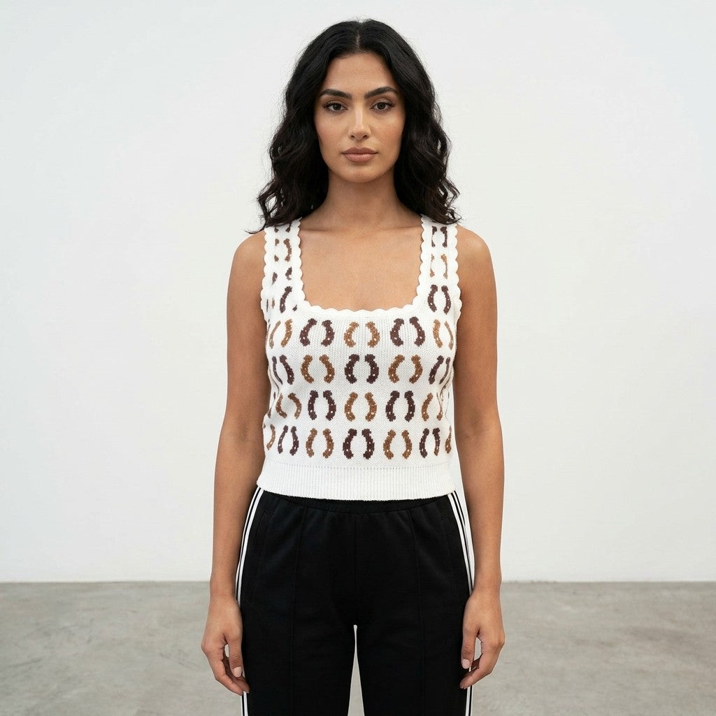 Woman with long hair, wearing a patterned white top and black pants, standing against a white wall.