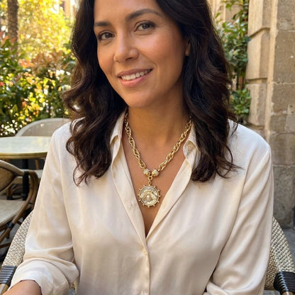 A woman smiles outdoors in a white blouse and a gold pendant necklace.