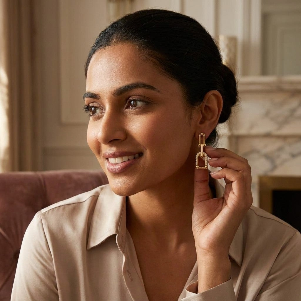 A smiling woman admires a gold dangling earring in a warmly lit room.