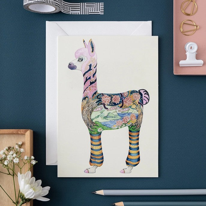 Colorful alpaca illustration on a card surrounded by stationery items.