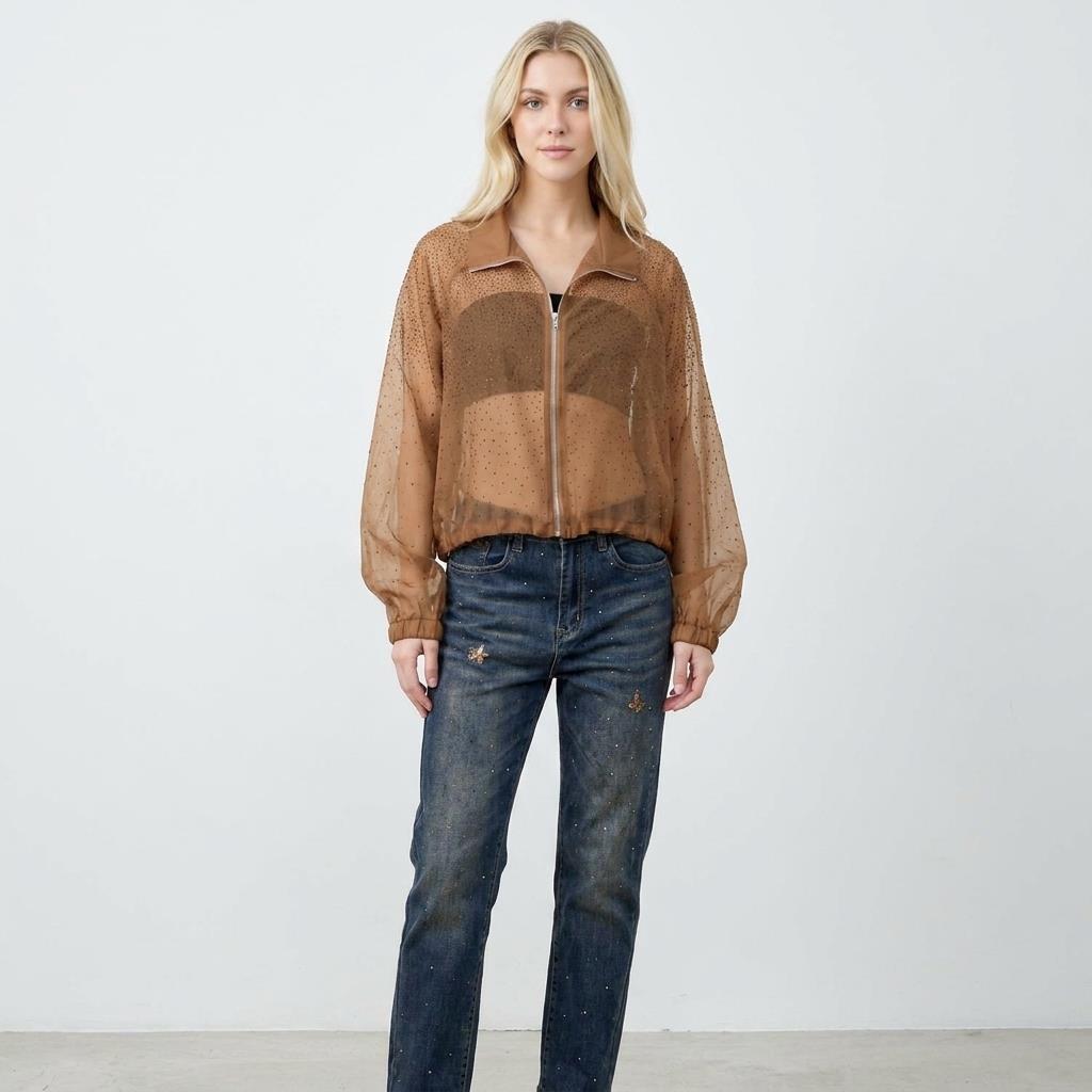 Woman in a sheer brown top and jeans, standing against a plain backdrop.
