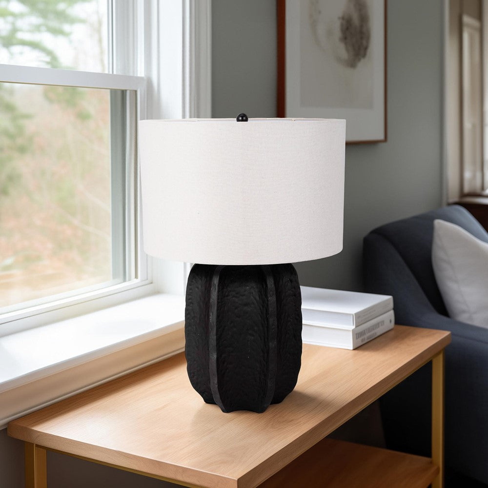 Black lamp with white shade on a wooden table by a window.