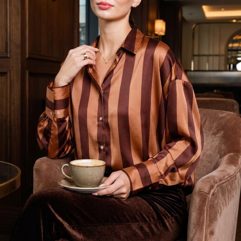 A woman in a striped satin blouse holds a cup of coffee in a cozy lounge.