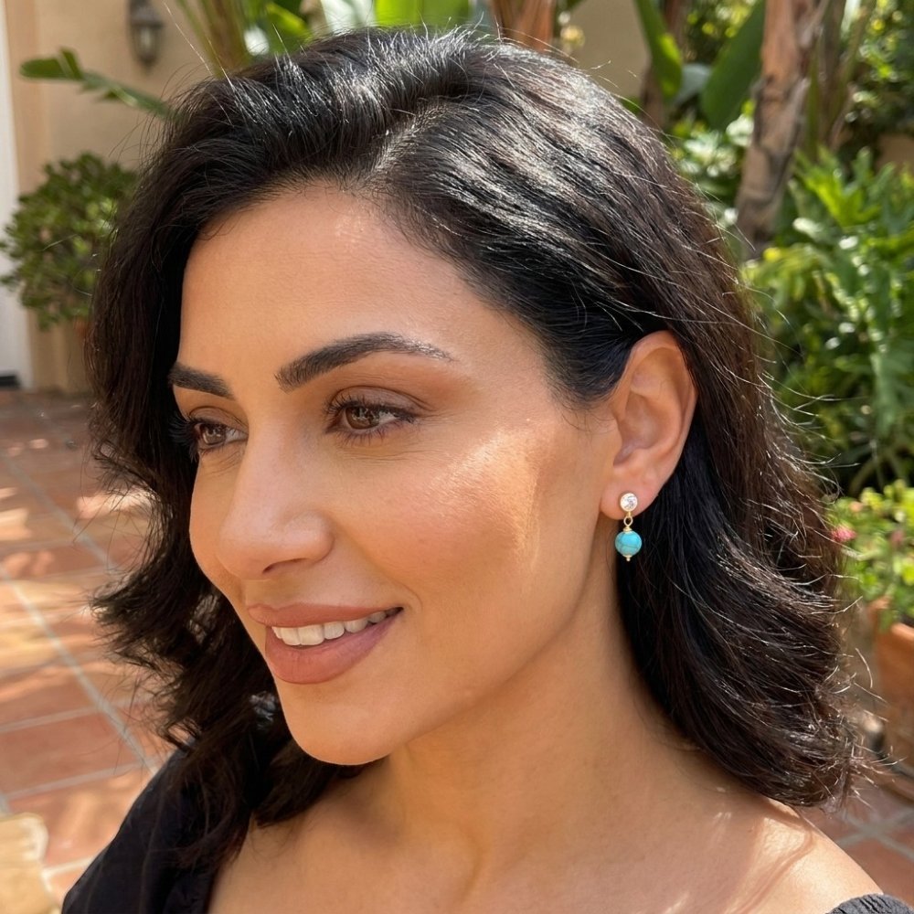 A smiling woman with dark hair and turquoise earrings poses in a sunny garden.