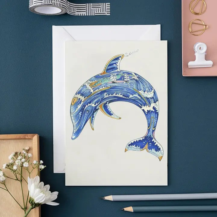 Illustrated dolphin card with floral decor and pencils on a blue surface.