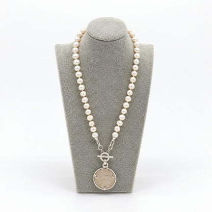 A pearl necklace with a silver toggle clasp and round pendant displayed on a gray stand.