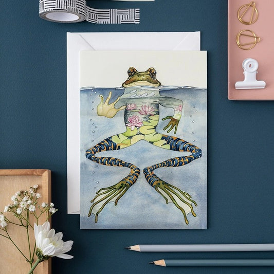 Artistic frog card on a desk with flowers and stationery.
