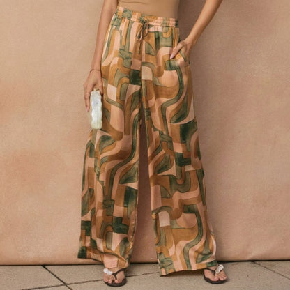 Woman in abstract pattern pants holding a bottle, standing against a beige wall.