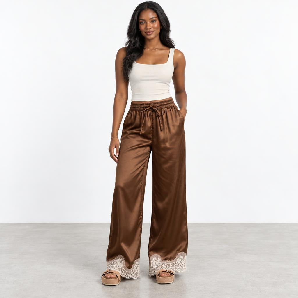 Woman in a white tank top and brown satin pants on a plain background.